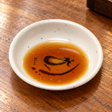 326-E Eggplant-pattern melamine sauce dish — Small amount of ponzu in lower half leaving part of motif visible through liquid, warm wooden restaurant table