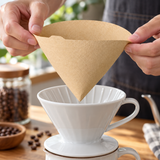 Natural HARIO V60 size 02 tabbed paper filter in a dry-filter loading setting. Dripper, server, coffee and surrounding scene accessories are illustrative, not included.