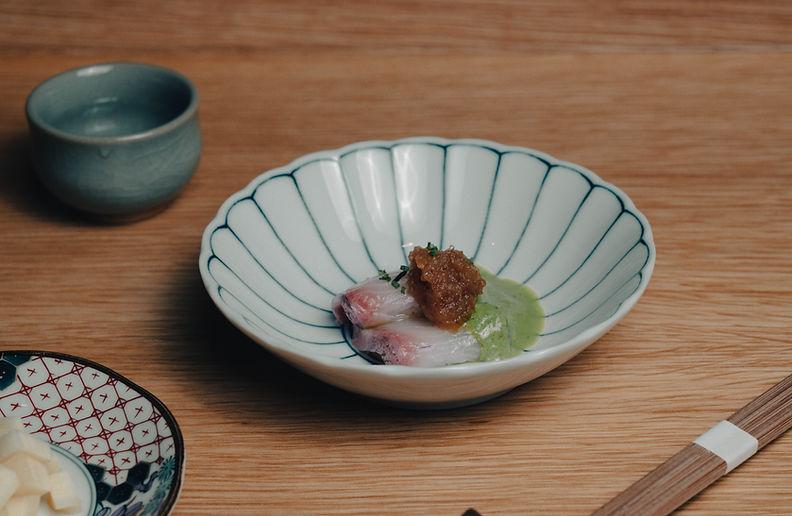 Fish presented in a blue-lined ceramic bowl, with a patterned side plate and chopsticks on a wooden table.