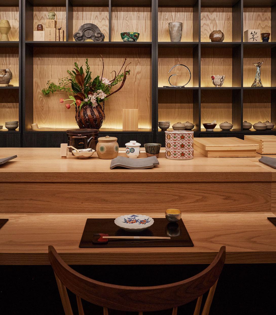 A Japanese counter setting with a patterned plate, dark tray, chopsticks and sake cup; varied servingware is organized behind the counter.