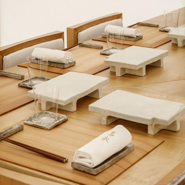 Repeated pale landing surfaces, glassware, chopsticks and small rests at a light-wood omakase counter.