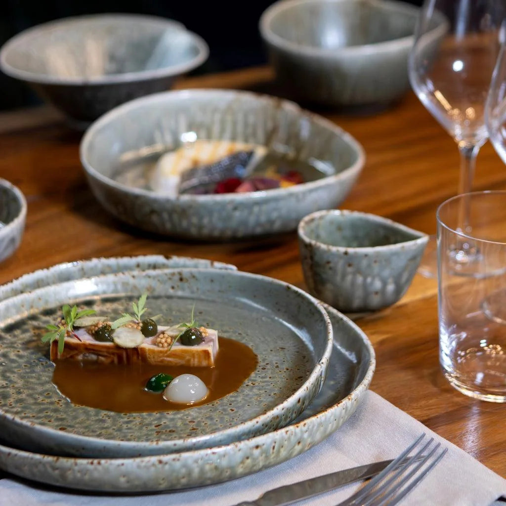 Pale mineral ceramic plates and bowls, a small cup and stemware arranged around a plated restaurant dish on a wooden table.