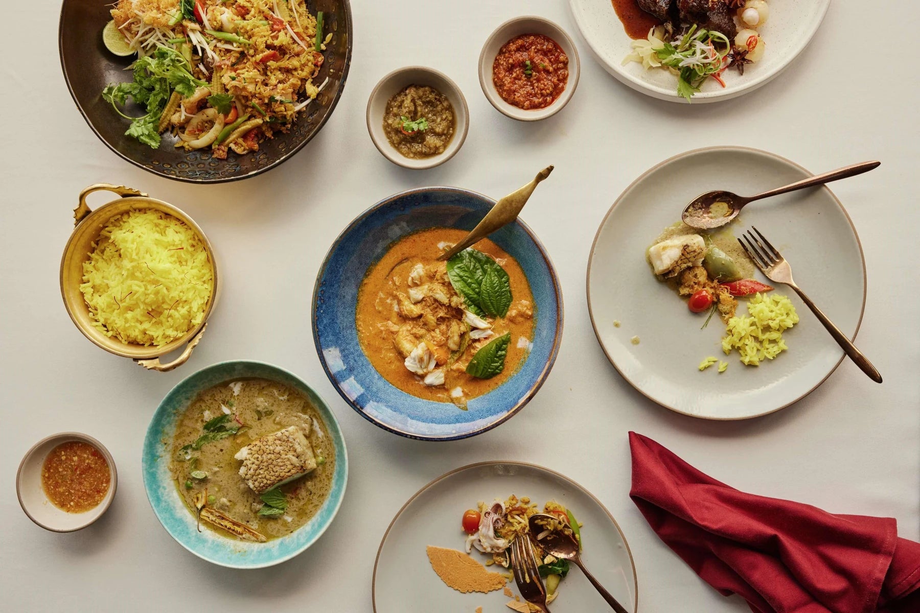 Overhead Thai meal with personal rice service, several shared curries and salads, small dipping dishes, forks and spoons.