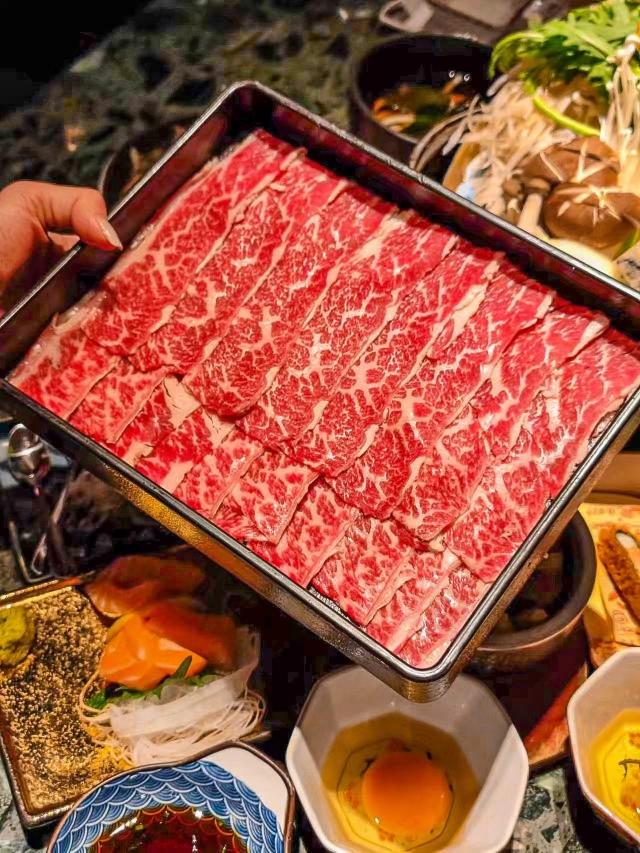 Sliced beef presented above a hot-pot setting with vegetables, an egg bowl and blue-patterned dipping ware.