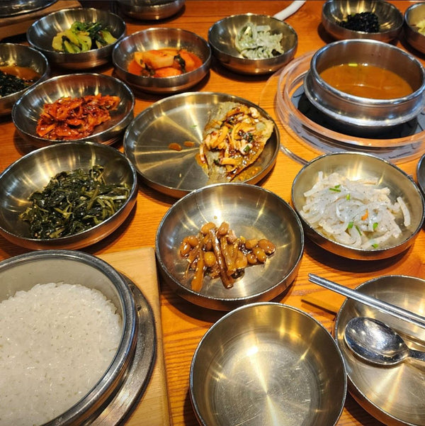 A Korean restaurant tabletop with repeated silver banchan dishes, rice service, metal utensils and a contrasting hot-food bowl.