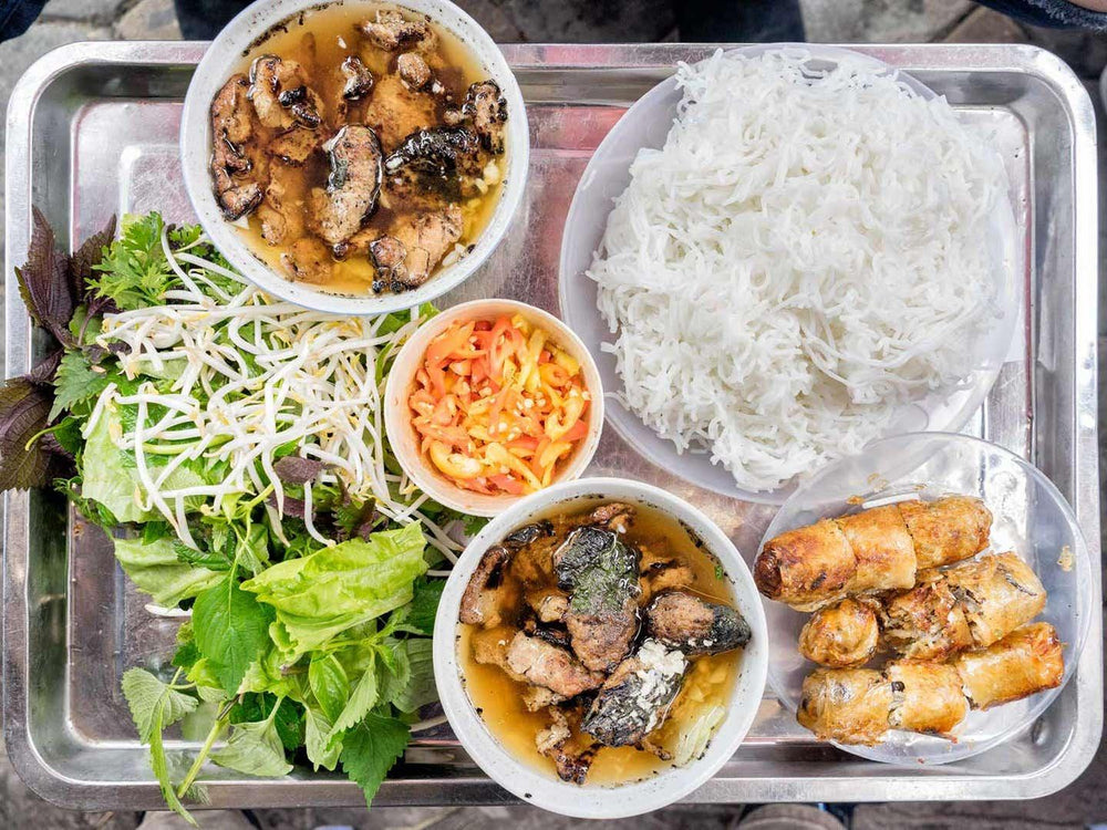 Bún chả service with grilled pork in dipping liquid, vermicelli, herbs, pickles and rolls in separate vessels.